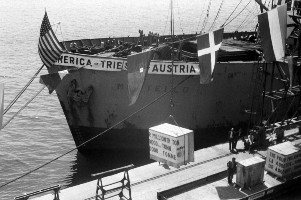 A ship unloads the four millionth ton. of E.R.P. aid. A banner with the names of States "America - Trieste - Austria" floats among the flags of the 18 Marshall Plan States and the U.S. flag.