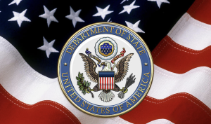 Il sigillo del Dipartimento di Stato USA in primo piano, dietro sventola la bandiera a stelle e strisce.