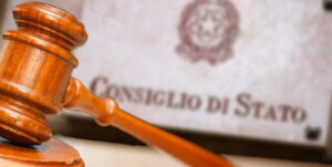 In primo piano un martelletto, sullo sfondo lo stemma della Repubblica Italiana e la scritta "Consiglio di Stato".